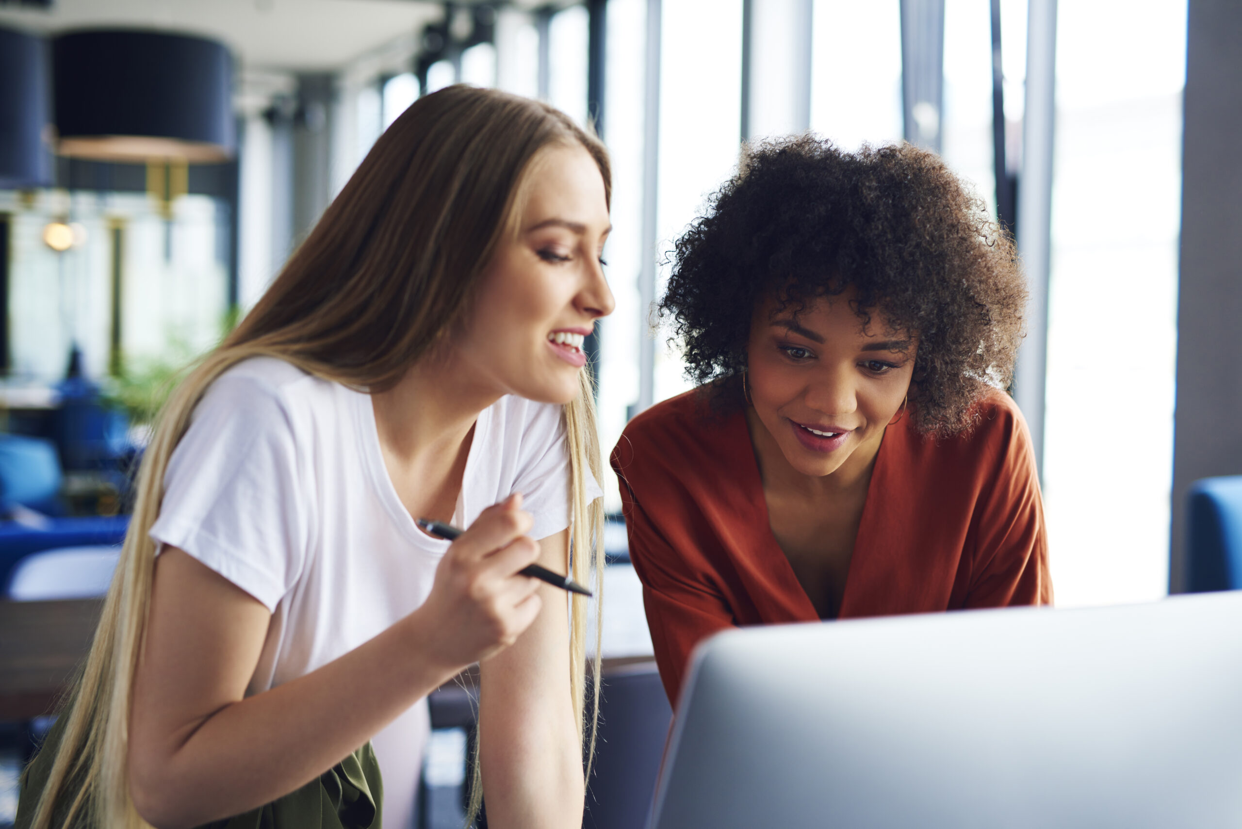 Deux femmes semblent discuter en regardant un écran d'oridinateur. La femme avec le t-shirt blanc semble suivre un accompagnement et prend des notes avec un stylo à la main.