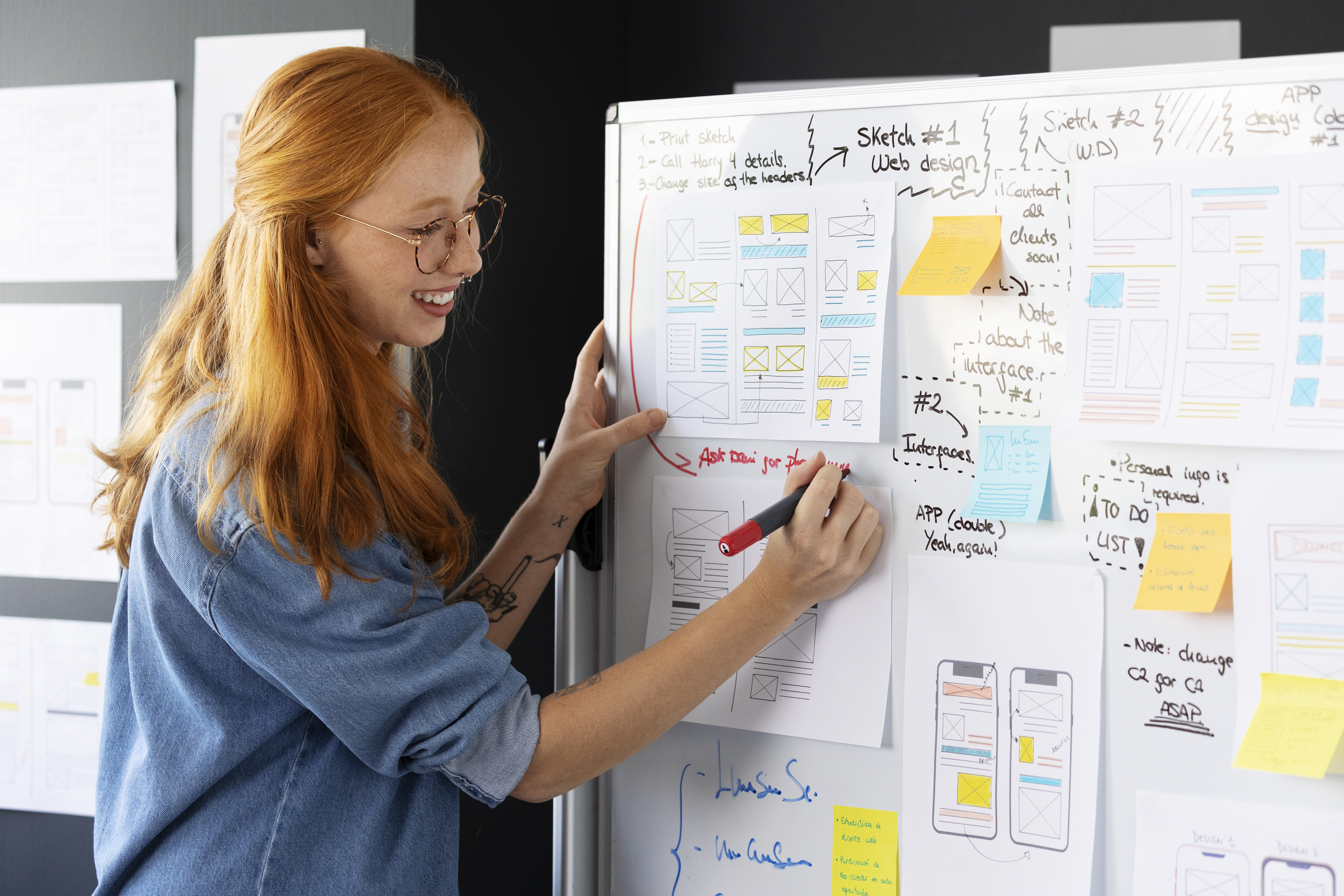 Une femme souriante qui porte des lunettes écrit au marqueur sur un tableau blanc. Sur celui-ci des séhémas, feuillets et post-its sont présents.